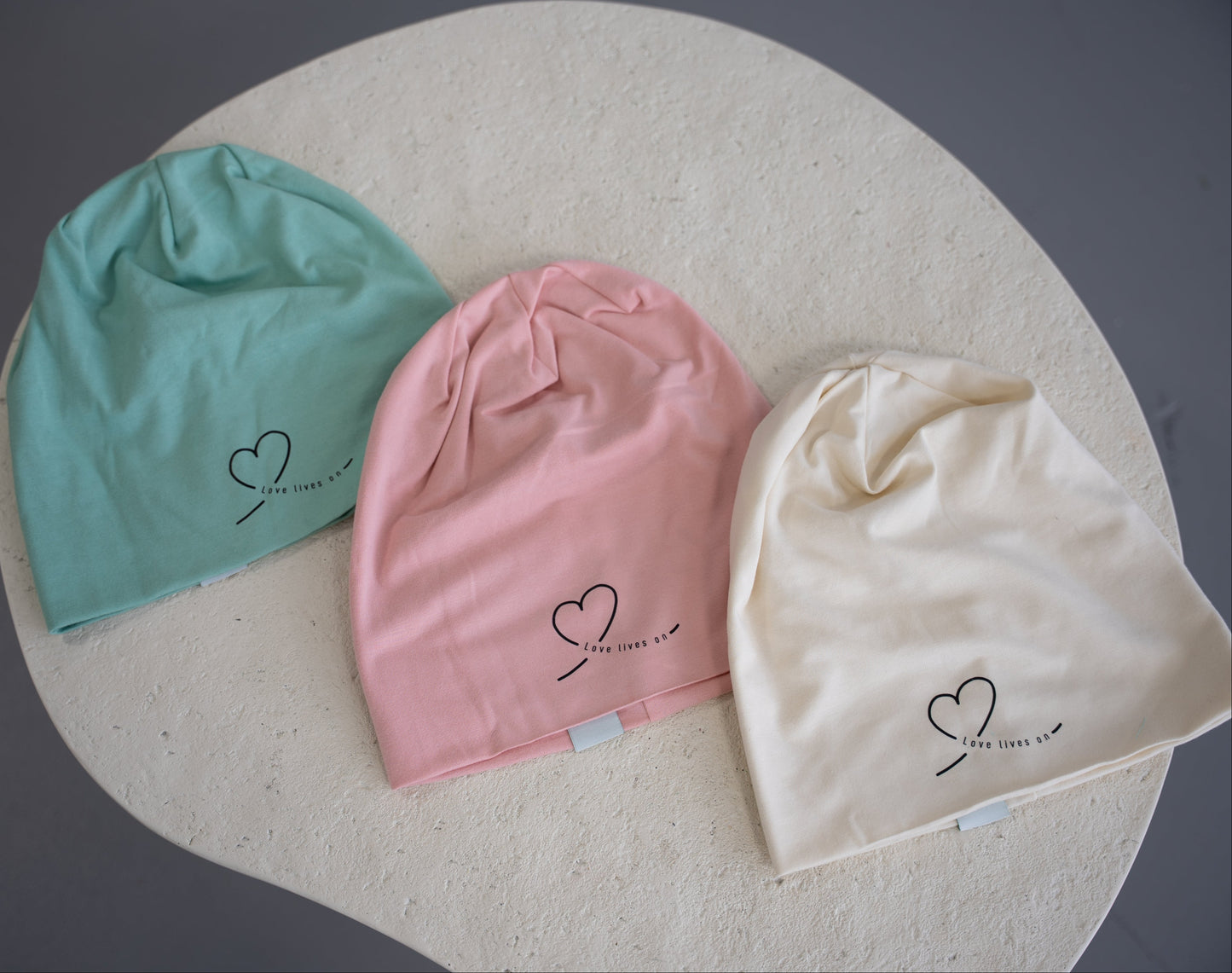 Three bamboo cotton beanie hats in pink, natural, and pistachio from HeartWear Co., featuring the “Love Lives On” logo.