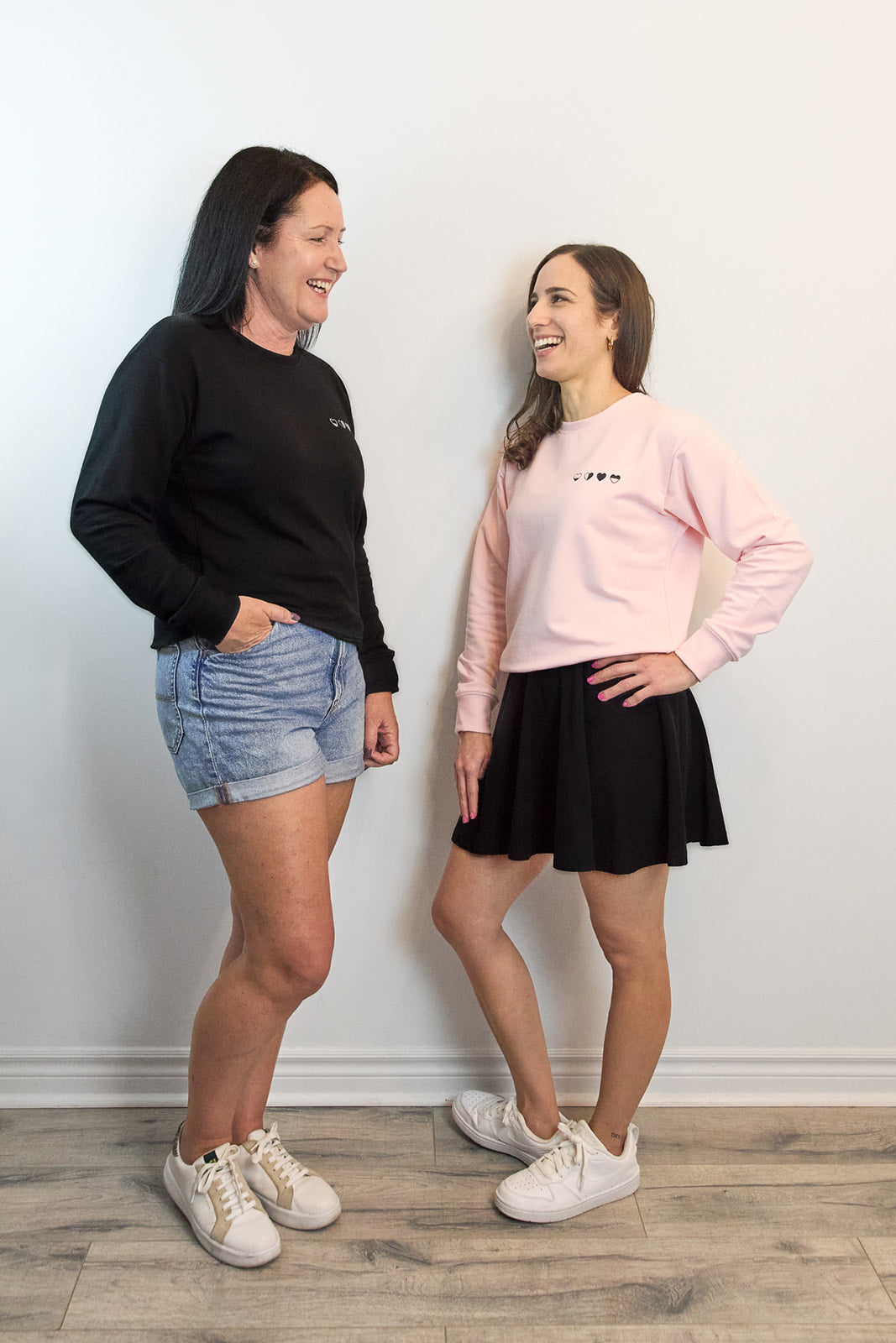 Two women in black and pink Heart Sweaters looking at each other—community, connection, and gentle support.