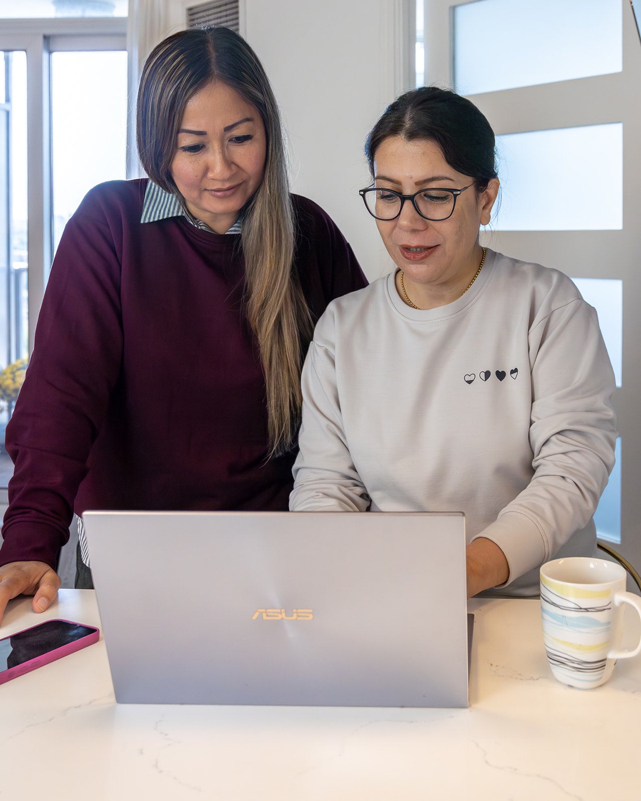 Two women in raspberry and stone Heart Sweaters at a laptop—comfort clothing for life’s busy, tender seasons.