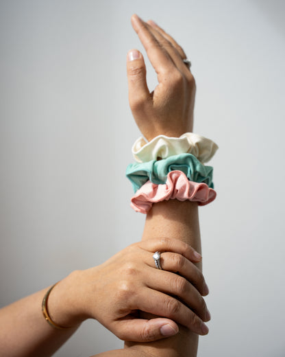 Hand holding three bamboo cotton scrunchies in pink, natural, and pistachio from HeartWear Co.’s Love Collection